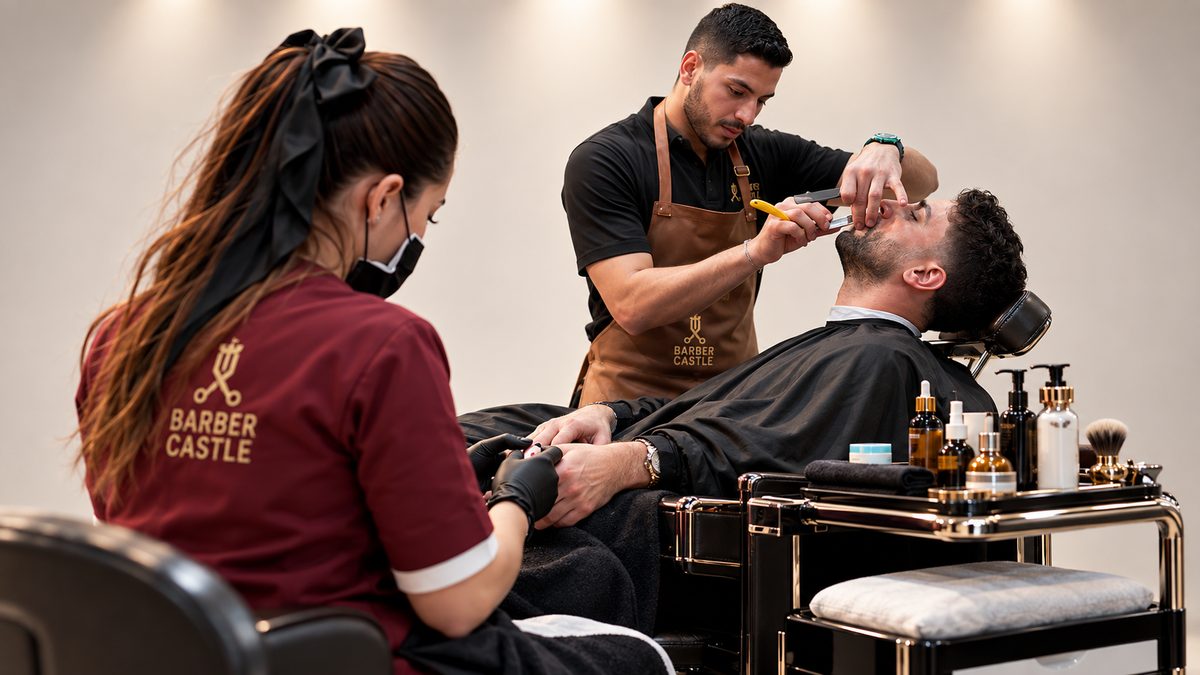 Close on an assistant in a Castle apron holding a client's hand while the barber works at his face behind.