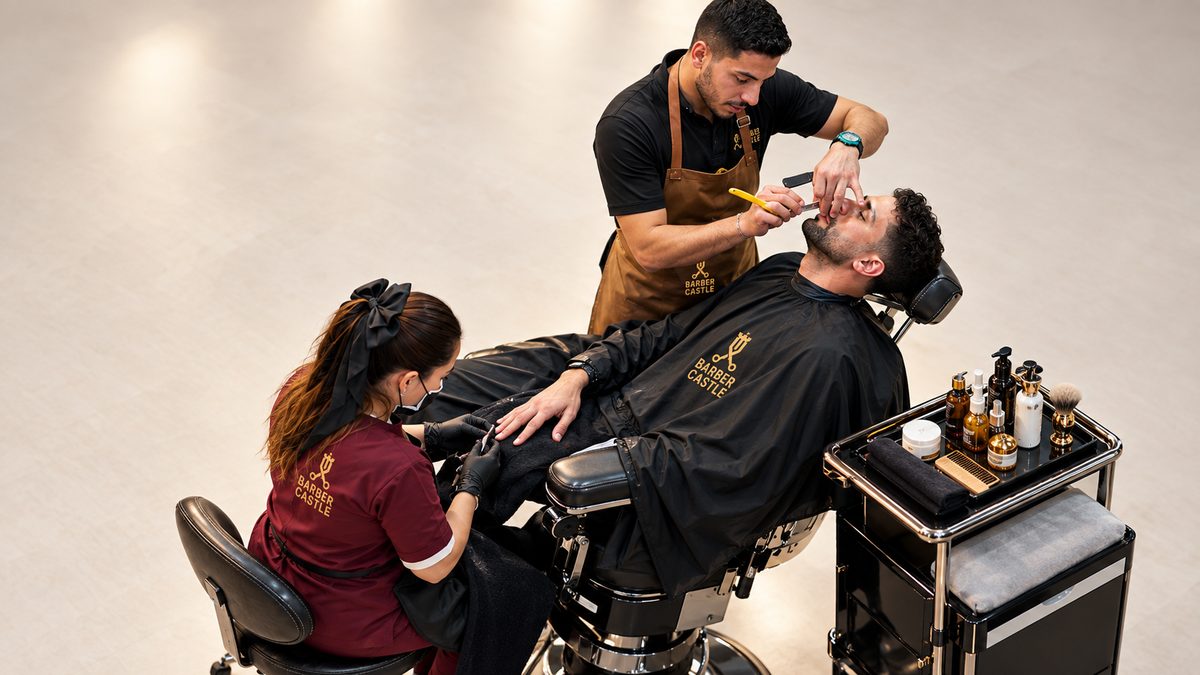 A barber drawing a straight razor down a client's cheek, the Castle crest printed on the client's cape, a brass trolley of tools beside the chair.