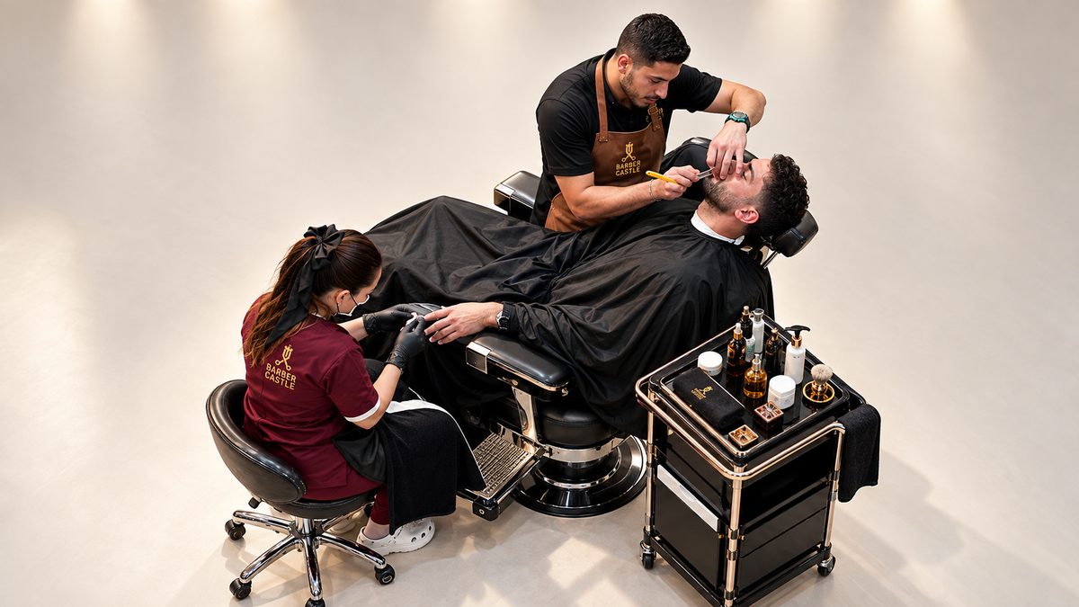 The whole station: the Castle chair, the client under a branded cape, the barber at the face and an assistant at his hands, beside a wheeled brass trolley.